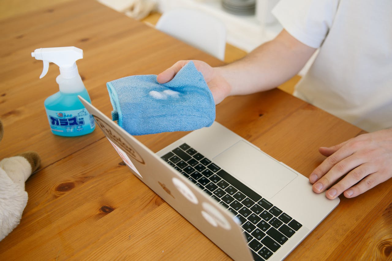 Person disinfecting laptop with spray and cloth, ensuring cleanliness and hygiene indoors.